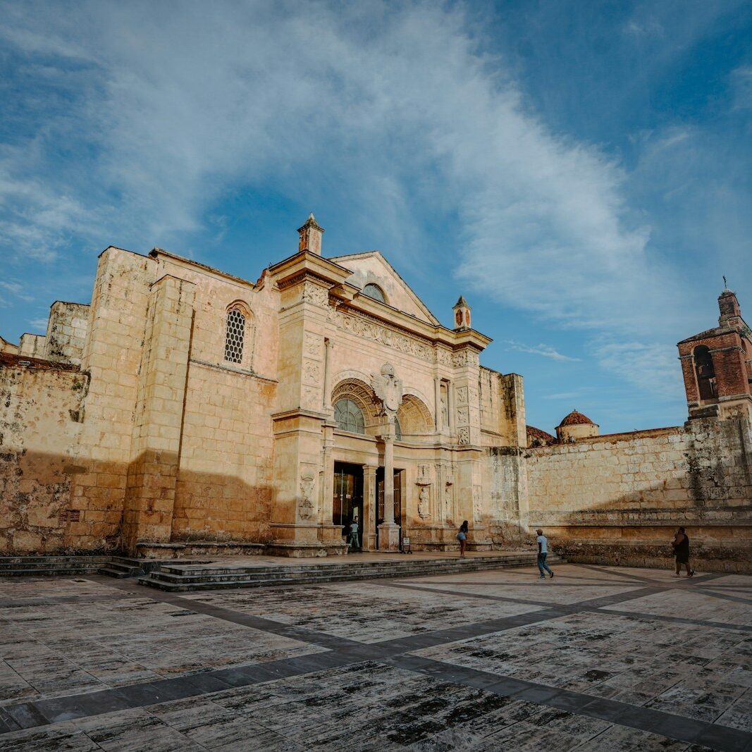 the Cathedral Primada de América, a historic building in Santo Domingo’s Zona Colonial. Its stone façade features simple yet elegant Gothic and Renaissance architectural details, with a central arched entrance and a few decorative carvings. The structure’s light-colored stone contrasts gently with the sky above. Cobblestone streets and a small plaza frame the cathedral, giving it a peaceful and timeless appearance.